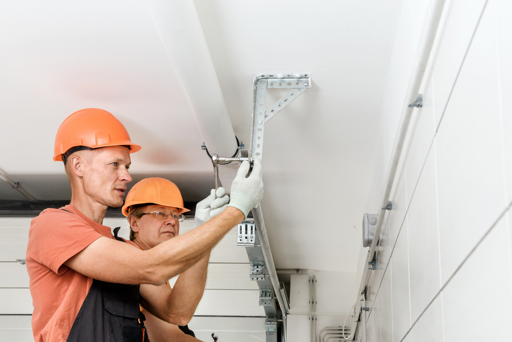 Two workers in orange hard hats tighten a bracket on a ceiling track system.